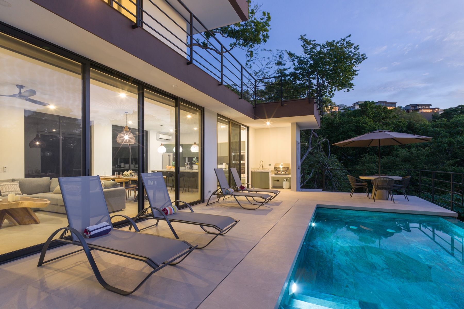 The pool terrace at dusk, with the lit living room and kitchen behind the glass doors
